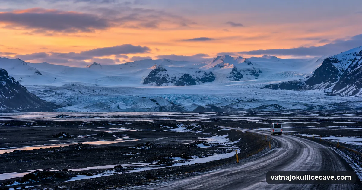 A road crossing a black sand plain at winter twilight, a glacier and snowy mountains on the horizon